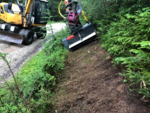 Ein moderner Bagger mit einer speziellen Fräse bearbeitet eine Böschung entlang eines Waldwegs. Die Maschine entfernt gezielt Bewuchs und bereitet den Boden für eine nachhaltige Pflege vor.